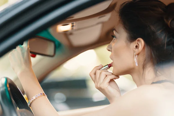 Mujer elegante joven mirando en el espejo de la vista del coche mientras se aplica maquillaje, lápiz labial en los labios — Foto de Stock