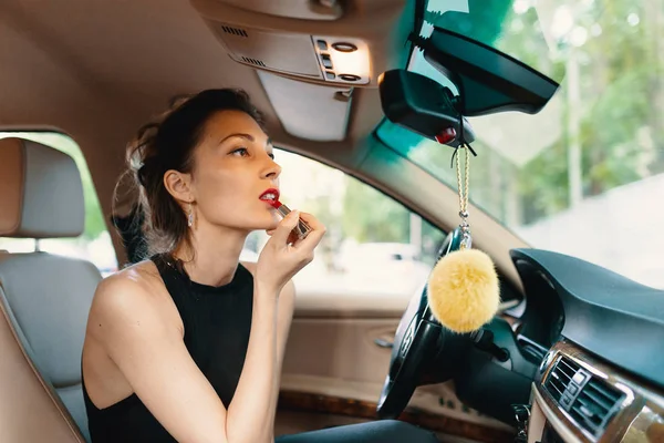 Jeune femme élégante regardant dans le rétroviseur de voiture tout en appliquant le maquillage, rouge à lèvres sur les lèvres — Photo