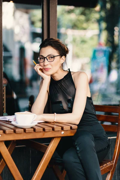 Mujer joven y elegante con gafas elegantes sentados en la mesa en la cafetería con una gran taza de café o café con leche — Foto de Stock