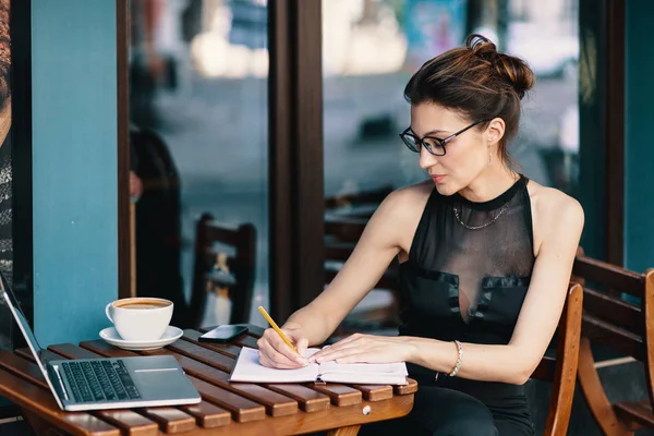 Femme d'affaires raffinée dans des lunettes, assise à la table dans un café travaillant à l'ordinateur portable — Photo