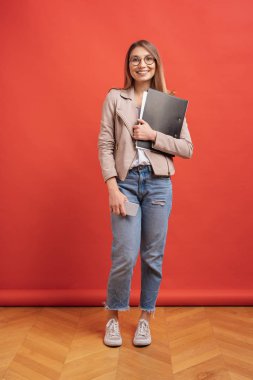 Genç gülümseyen öğrenci veya kırmızı arka plan üzerinde bir klasör ile duran gözlük stajyer.