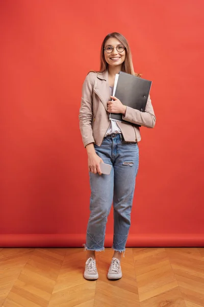 Joven estudiante sonriente o pasante en gafas de pie con una carpeta sobre fondo rojo . — Foto de Stock