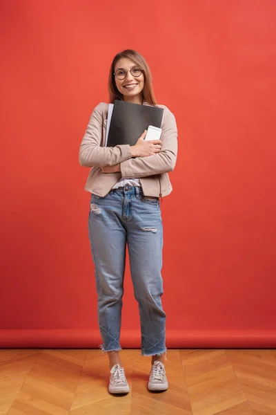 Joven estudiante sonriente o pasante en gafas de pie con una carpeta sobre fondo rojo . — Foto de Stock