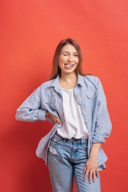 Komik, şirin kız kırmızı bir arka plan üzerinde göz kırpıyor ve dilini gösteren izole eğleniyor.