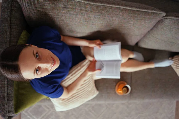 Atractiva mujer positiva leyendo un libro relajándose en el sofá —  Fotos de Stock