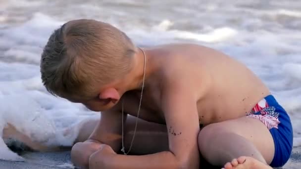 Niedliches kleines Baby, das am Strand sitzt und die Wellen genießt, die mit dem Sand spielen. Zeitlupe — Stockvideo