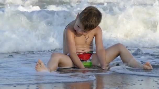 Słodkie małe dziecko siedzi na plaży i cieszyć się fale, grając z piasku. Zwolnionym tempie — Wideo stockowe