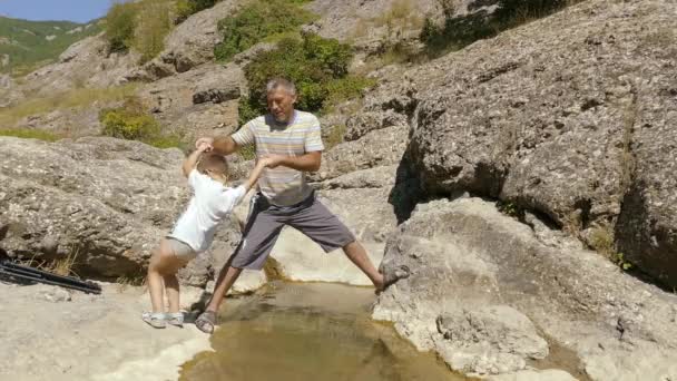 Père aide un petit fils avec des lunettes à traverser le ruisseau de montagne . — Video