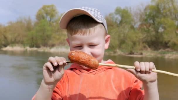 The kid eats the sausage with his teeth, strung on the willow twig with pleasure. — Stock Video