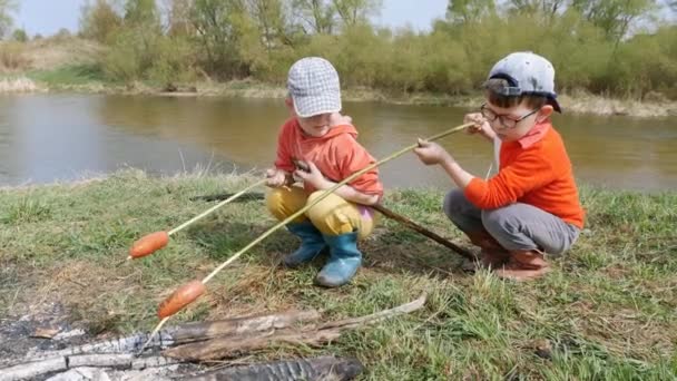 Два маленьких мальчика пекут сосиски на углях на берегу быстрой реки. . — стоковое видео