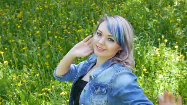 Young blonde girl with professional makeup and hairstyle blue hair in a black dress and wearing a denim jacket dancing moves in front of the camera in the green grove. The camera is in motion. — Stock Video