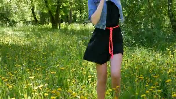 Jeune fille blonde avec un maquillage professionnel et coiffure cheveux bleus dans une robe noire et portant une veste en denim danse se déplace devant la caméra dans le bosquet vert. La caméra est en mouvement . — Video