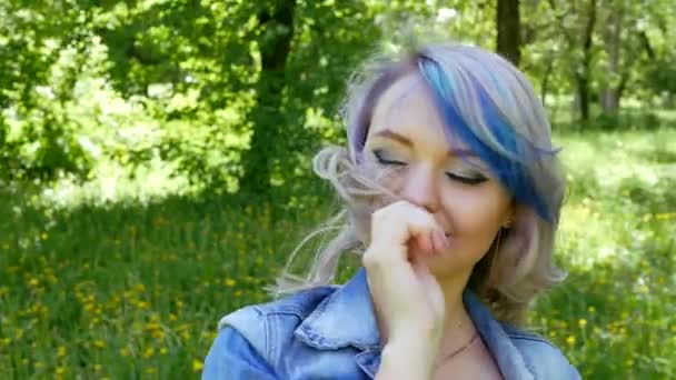 Young blonde girl with professional makeup and hairstyle blue hair in a black dress and wearing a denim jacket dancing moves in front of the camera in the green grove. The camera is in motion. — Stock Video