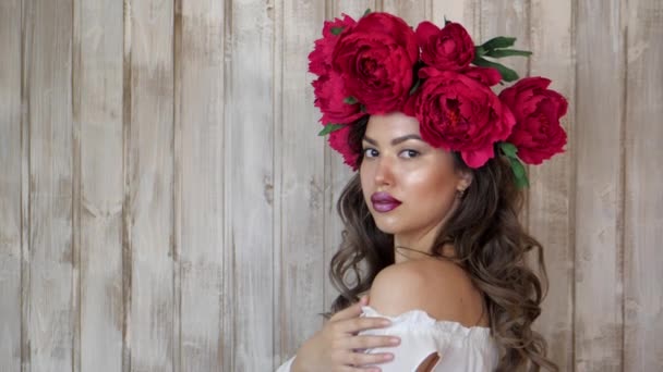 Chica posando delante de la cámara. mujer joven en una corona de peonías escarlata en su cabeza, pelo largo oscuro rizado desciende sobre los hombros poco profundos . — Vídeos de Stock