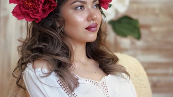 Girl posing in front of camera. young woman in a wreath of scarlet peonies on her head, dark long curly hair descends on the shallow shoulders. — Stock Video