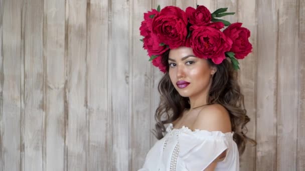 Girl posing in front of camera. young woman in a wreath of scarlet peonies on her head, dark long curly hair descends on the shallow shoulders. — Stock Video