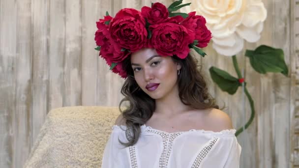 Chica posando delante de la cámara. mujer joven en una corona de peonías escarlata en su cabeza, pelo largo oscuro rizado desciende sobre los hombros poco profundos . — Vídeos de Stock