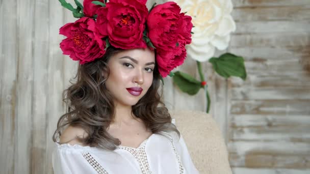 Girl posing in front of camera. young woman in a wreath of scarlet peonies on her head, dark long curly hair descends on the shallow shoulders. — Stock Video