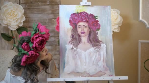 Artista disegna un ritratto dalla natura. L'artista disegna un ritratto dalla natura. Fuoco flottante della telecamera, telecamera in movimento. Bella modella, con una corona di peonie scarlatte in testa, in posa seduta in — Video Stock