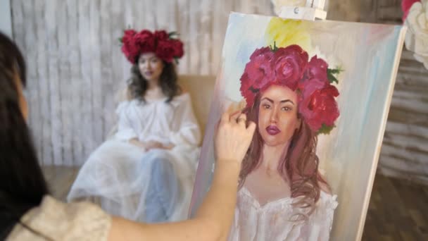 Artista disegna un ritratto dalla natura. L'artista disegna un ritratto dalla natura. Fuoco flottante della telecamera, telecamera in movimento. Bella modella, con una corona di peonie scarlatte in testa, in posa seduta in — Video Stock