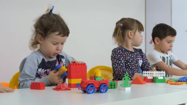 Niños desarrollando una sala de juegos. Emociones de los niños pequeños durante las clases de entretenimiento. Niños y niñas están jugando con bloques de madera sentados a la mesa . — Vídeo de stock