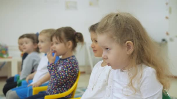 Kinderen ontwikkelen een spelletjeskamer. De emoties van jonge kinderen tijdens het onderhoudend klassen. Kinderen zitten op stoelen in een rij. Het kind bouwt een vrolijke expressie. Grimaces en smeyotsya plezier. — Stockvideo