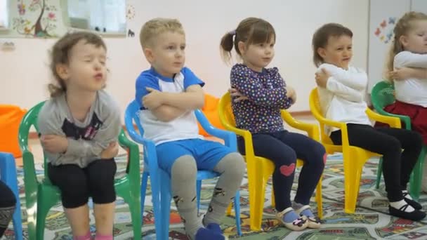 Niños desarrollando una sala de juegos. Emociones de los niños pequeños durante las clases de entretenimiento. Los niños se sientan en sillas seguidas. El niño se abraza con las manos y luego las levanta a un lado . — Vídeo de stock