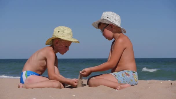 Odpočiň si na moři s dětmi. Písek se vylévá z dlaní rukou do dlaní dítěte. Dva kluci v kloboucích s okrajem na pobřeží. — Stock video