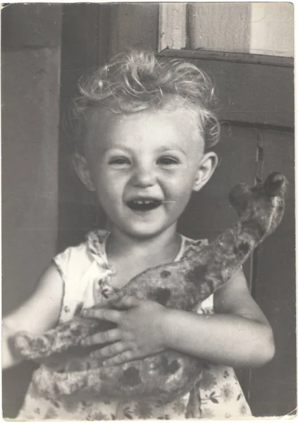 História nas fotos: Menina com cabelo encaracolado com um brinquedo em suas mãos. Uma criança tem uma girafa. Retrato, 1983, URSS . Imagem De Stock