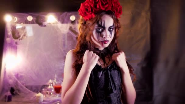 Maquillaje fácil de Halloween. La chica con la foto en la cara. La novia de los demonios con una corona de flores rojas en la cabeza. La mujer lleva un vestido de corsé negro y medias negras. La chica haciendo — Vídeos de Stock