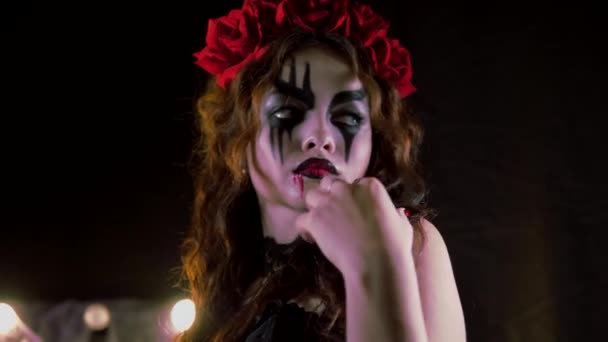 Gemakkelijke Halloween make-up. Het meisje met de foto op haar gezicht. De duivelse bruid met een krans van rode bloemen op haar hoofd. Het meisje maakt goo-goo ogen en toont zichzelf. — Stockvideo