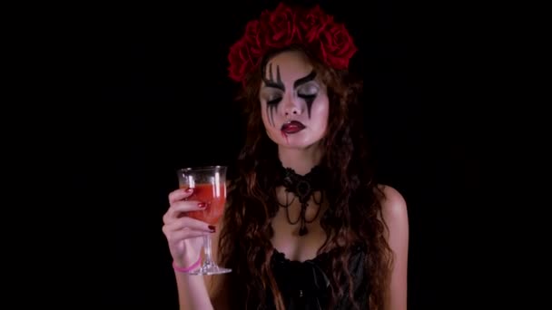 Lehký Halloweenský makeup. Dívka s obrázkem na tváři. Ďábelská nevěsta s věnečkem červených květin na hlavě. Žena pije ze sklenice červeného nápoje a nabízí divákovi pití. — Stock video