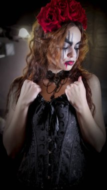 Kolay Cadılar Bayramı makyajı. Yüzünde resim olan kız. Şeytanın gelini kafasında kırmızı çiçek çelengiyle. Kadın siyah bir korse ve siyah çorap giyiyor..