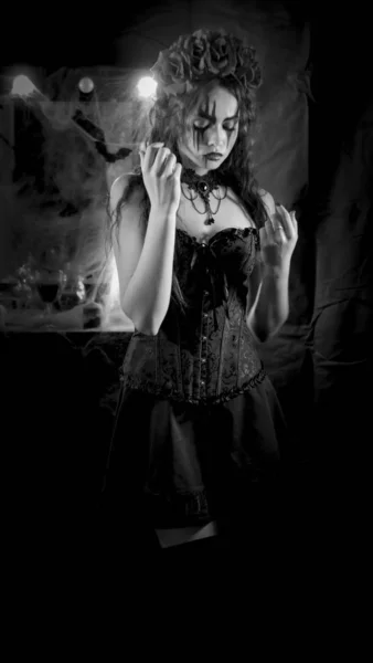 Maquillage Halloween facile. La fille avec la photo sur le visage. La mariée du diable avec une couronne de fleurs rouges sur la tête. La femme porte une robe corset noire et des bas noirs . — Photo