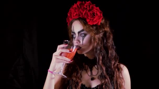 Εύκολη Απόκριες μακιγιάζ. Το κορίτσι με τη φωτογραφία στο πρόσωπό της. Η διαβολική νύφη με ένα στεφάνι από κόκκινα λουλούδια στο κεφάλι της. Γυναίκα πίνει από ένα ποτήρι κόκκινο ποτό προσφέροντας να πιει στον θεατή. — Αρχείο Βίντεο