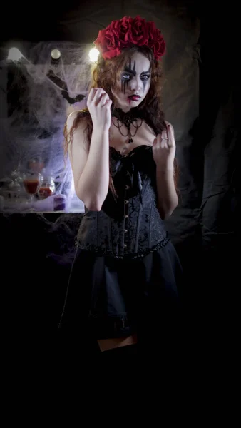 Легкий макияж на Хэллоуин. Девушка с фотографией на лице. Невеста дьявола с венком из красных цветов на голове. На женщине чёрное корсетное платье и чёрные чулки. . — стоковое фото