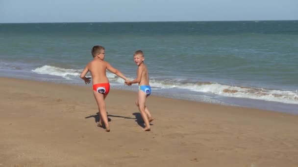 I bambini corrono lungo la costa. Onde lavarsi sulla spiaggia. Ragazzi che si tengono per mano correndo lungo la spiaggia. Il ragazzo con gli occhiali sul resto del mare. Il ragazzo corre e torna indietro . — Video Stock