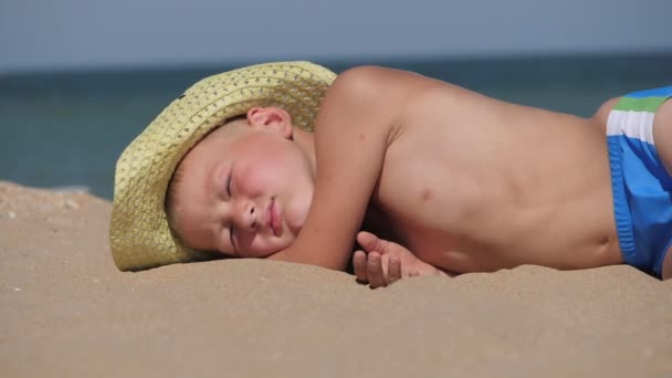 El chico yace en la arena dorada. El niño lleva un sombrero de paja con un ala grande. El niño duerme en la playa . — Vídeos de Stock