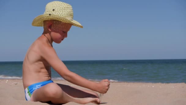 Мальчик сидит на золотом песке. Ребенок носит соломенную шляпу с большими краями. Ребенок играет на песке на пляже. Светло-зеленая шляпа развивается на ветру . — стоковое видео