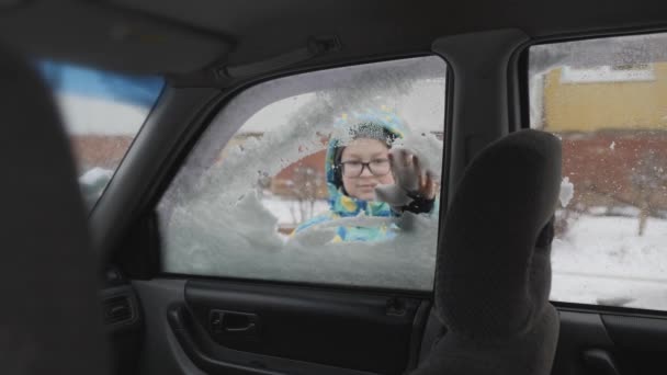 Die Seitenscheibe des Autos vom Schnee befreien. Eine Kinderhand reinigt die Autoscheibe. Der Junge lächelt süß, während er das Glas putzt. — Stockvideo