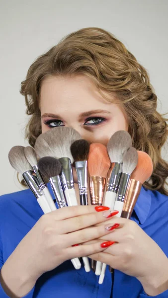 Young blonde girl hides her lips behind makeup brushes. The face is hidden behind curls of hair. Clearly expressed emotions on the womans face.