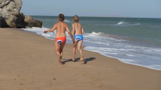 As crianças correm ao longo da costa. Ondas se lavam sobre a praia. Rapazes de mãos dadas a correr pela praia. O miúdo dos óculos no resto do mar. O menino corre e volta . — Vídeo de Stock