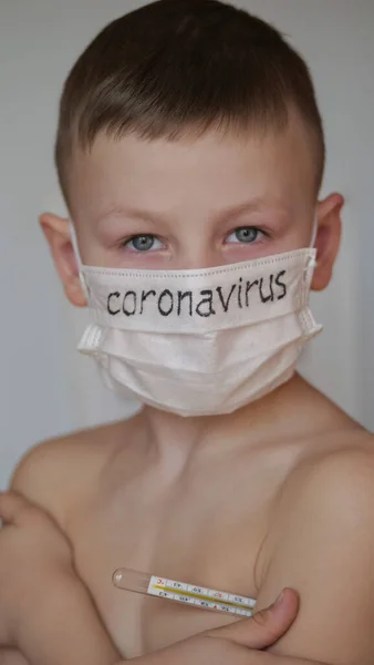 Медицинская маска на лице ребенка. На белой маске чёрная надпись Коронавирус. Блондин осторожен с вирусом. Термометр ртути под рукой у детей . — стоковое фото