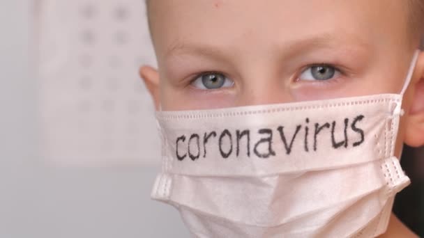 Máscara médica en la cara de los niños. En la máscara blanca hay una inscripción negra Coronavirus. El chico rubio tiene cuidado con el virus. Termómetro de mercurio bajo la mano de los niños . — Vídeos de Stock