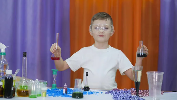 Kind hält Kolben mit farbiger Flüssigkeit in der Hand. Junge mit durchsichtiger Schutzbrille im Gesicht. Chemische Experimente für Kinder. Lustige Experimente für Kinder. Kinder haben Spaß und lernen. Hell — Stockfoto