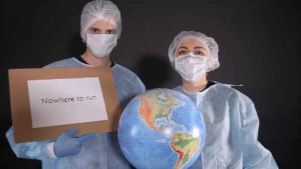 Man and a woman in protective clothing hold signs with the words NOWHERE TO RUN and QUARANTINE. The girl is holding a model of the earth, a globe. Protective mask on the face of people. On the white — Stock Video