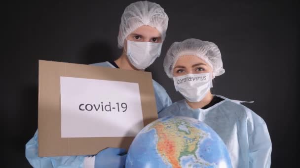 Мужчина и женщина в защитной одежде держат знаки со словами covid-19 и QUARANTINE. Девушка держит в руках модель Земли, глобус. На белой маске черная надпись Коронавирус . — стоковое видео