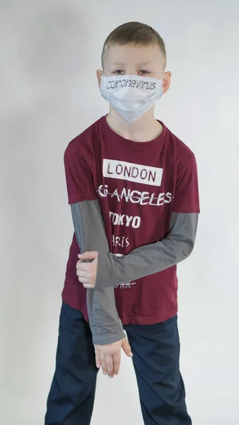 Boy si alza in piedi indossando una t-shirt con le parole: Parigi, Los Angeles, Tokyo, New York, Berlino. Sulla maschera bianca è un'iscrizione nera Coronavirus . — Foto Stock