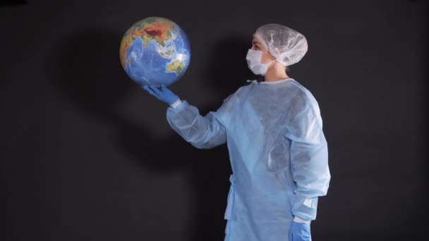 Meisje in lichte beschermende kleding. Op het gezicht van de persoon is een medisch masker. Haar haar is bedekt met een medisch kapje. Een jonge vrouw houdt een model van de planeet aarde vast.. — Stockvideo