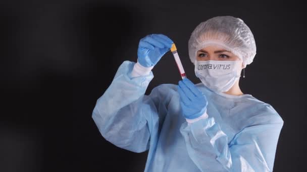 Girl examina un tubo de ensayo con biomaterial. Una joven con ropa protectora. En la cara de una máscara médica protectora. Las manos están vestidas con guantes de látex azul. En la máscara blanca-negro — Vídeos de Stock